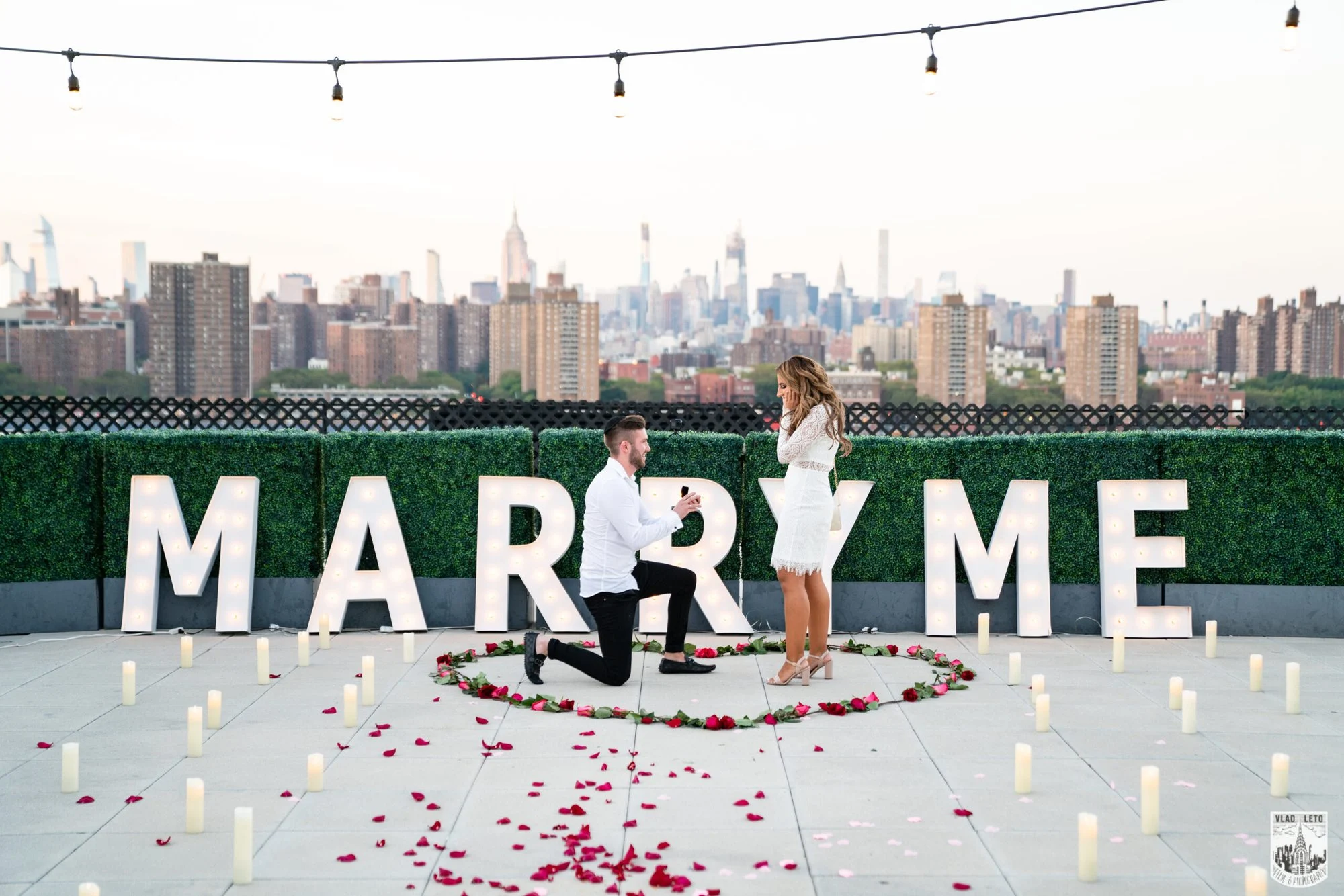 Man on one knee proposing inside a rose-petal heart and candles in front of white MARRY ME letters and a skyline view