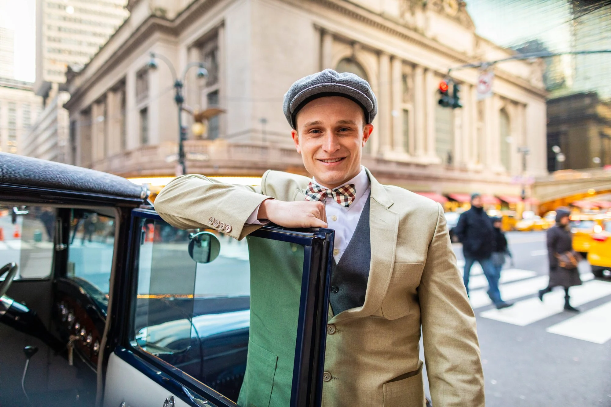 Man in a tweed suit, bow tie and flat cap leaning on a vintage car door with a grand stone building behind
