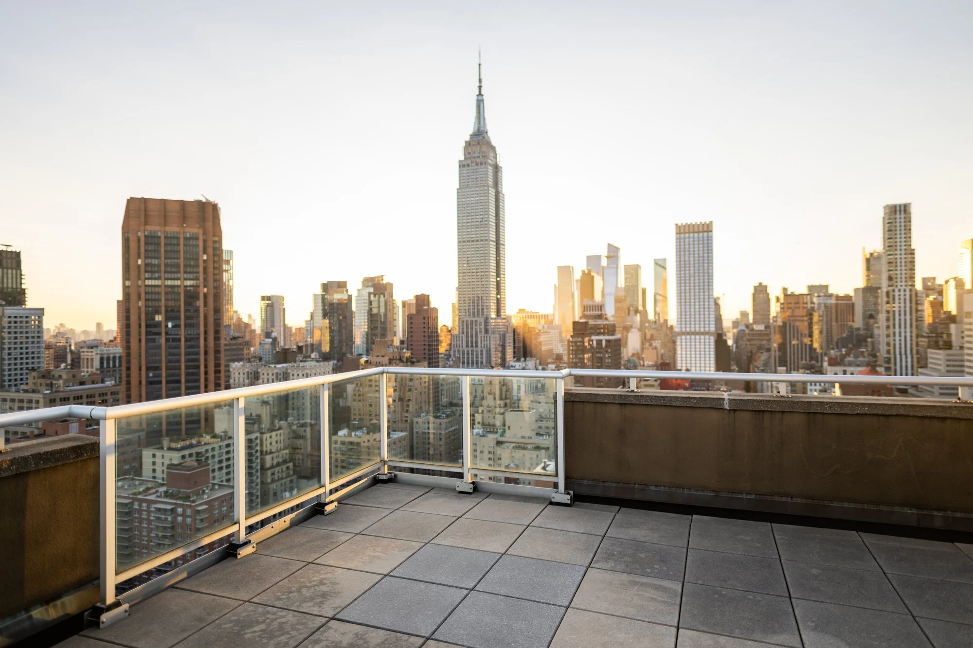 Empty glass-railed rooftop terrace in Manhattan with the Empire State Building and skyline glowing at golden hour