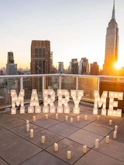 Lit MARRY ME marquee letters and candles on a Manhattan rooftop at sunset with the Empire State Building behind