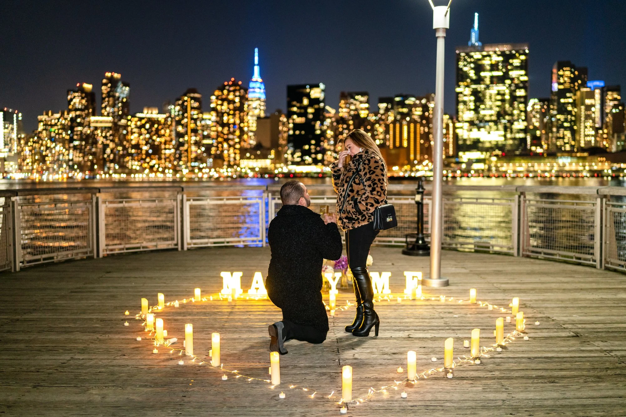 Man kneeling on a wooden pier inside a candle heart with MARRY ME letters as his partner reacts, skyline lit behind them