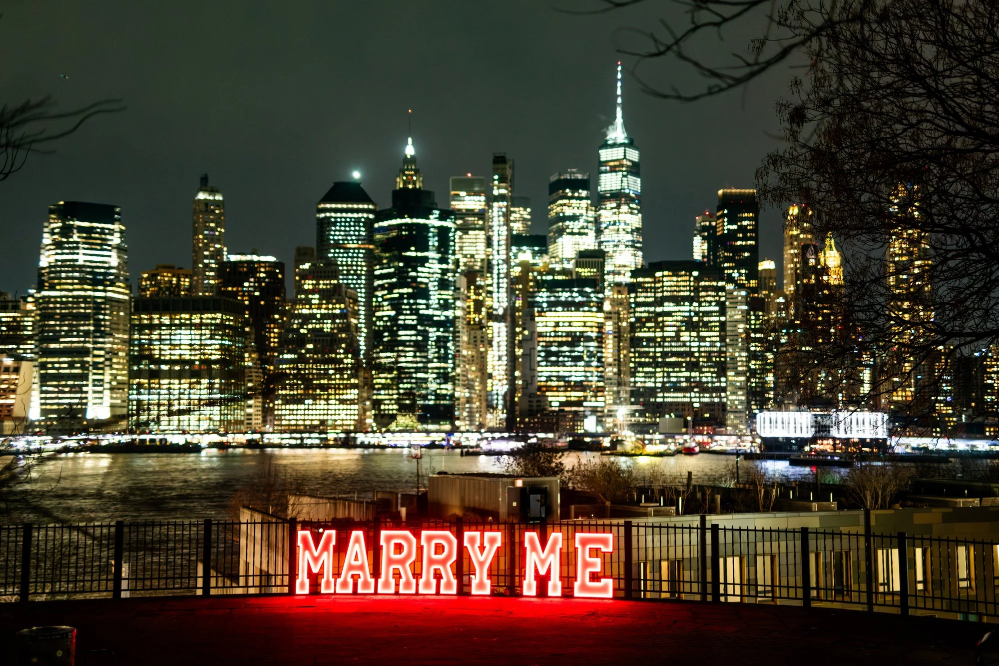 Glowing red MARRY ME letters by a waterfront railing with the lit Lower Manhattan skyline and One World Trade at night
