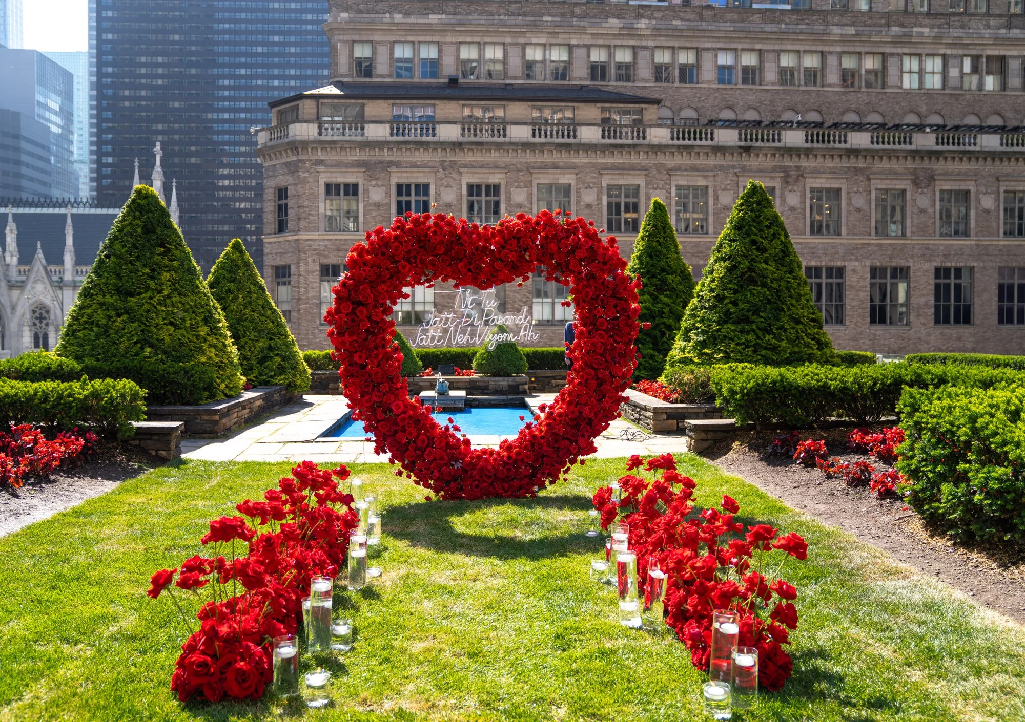 Giant red rose heart arch with rose clusters and candles on a green lawn, with a reflecting pool and stone buildings behind