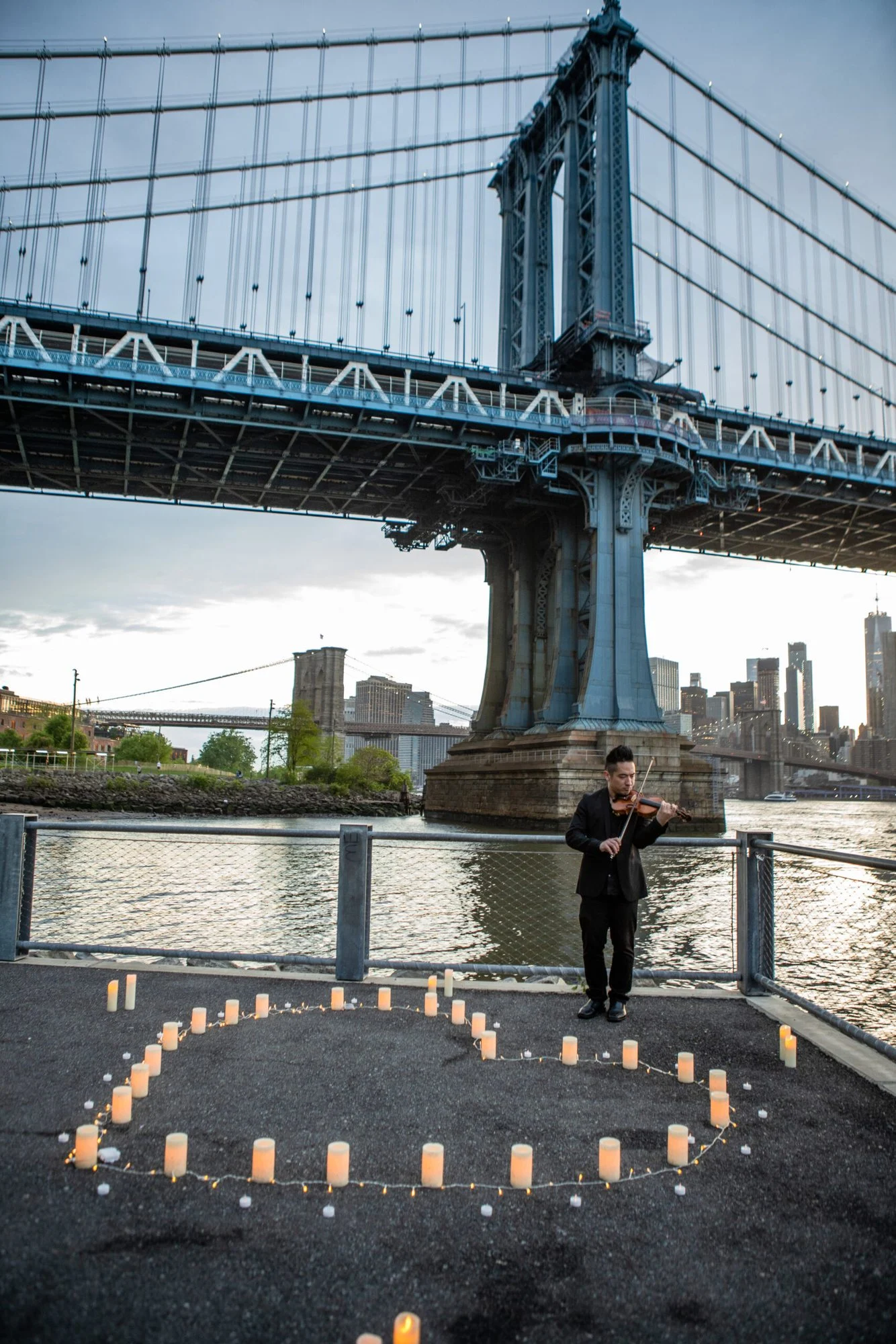 Violinist standing by the water beside a heart of candles under a Manhattan Bridge tower