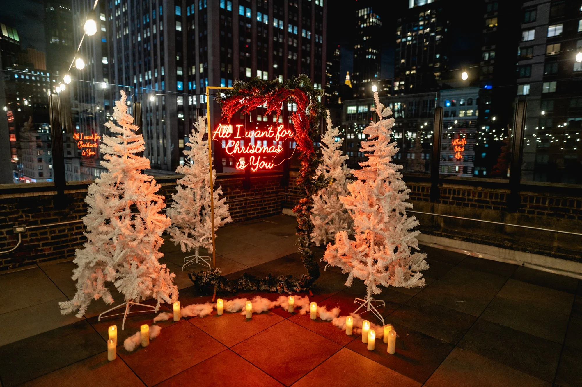 Two white flocked Christmas trees and candles flank a red neon Christmas sign on a rooftop with string lights