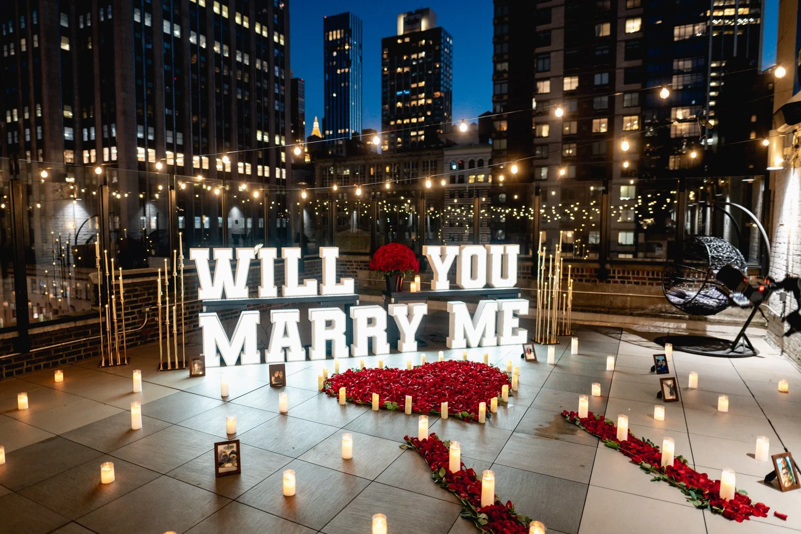 Lit WILL YOU MARRY ME letters, a rose-petal heart and candles under string lights on a Manhattan rooftop at night